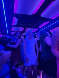 Friends dancing inside a neon-lit party bus interior with purple and blue LED ceiling panels; person in a white dress smiling at center.