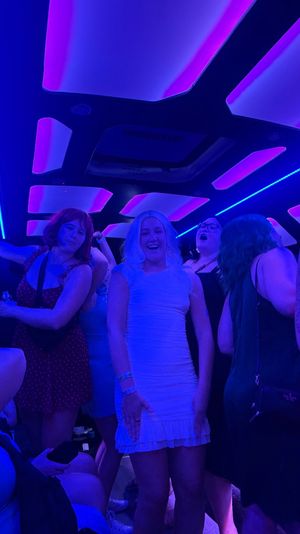 Friends dancing inside a neon-lit party bus interior with purple and blue LED ceiling panels; person in a white dress smiling at center.