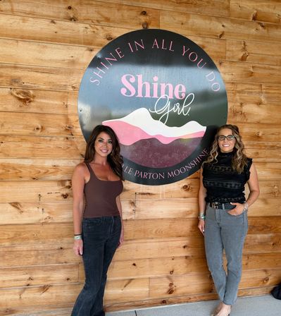 Two smiling women standing beside a large round pink-and-black mountain logo sign mounted on a rustic wood plank wall, casual outfits and relaxed photo-op vibe.
