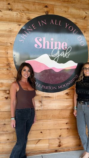 Two smiling women standing beside a large round pink-and-black mountain logo sign mounted on a rustic wood plank wall, casual outfits and relaxed photo-op vibe.
