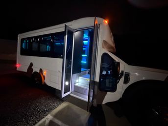 White shuttle-style party bus parked at night in a lot, open folding door revealing lit steps and blue LED interior lighting with shadowed silhouettes near the rear wheel.
