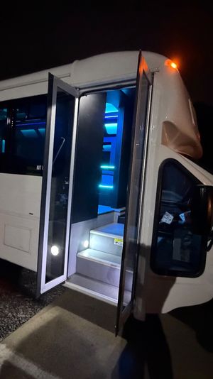 White shuttle-style party bus parked at night in a lot, open folding door revealing lit steps and blue LED interior lighting with shadowed silhouettes near the rear wheel.
