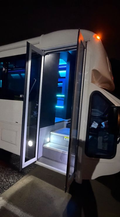 White shuttle-style party bus parked at night in a lot, open folding door revealing lit steps and blue LED interior lighting with shadowed silhouettes near the rear wheel.