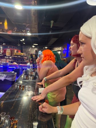 Group of people wearing colorful wigs (white, red, orange, green) leaning on a glossy bar tasting counter lined with shot glasses under blue neon lights in a lively indoor bar.