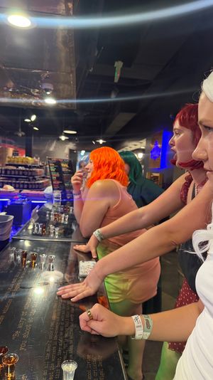 Group of people wearing colorful wigs (white, red, orange, green) leaning on a glossy bar tasting counter lined with shot glasses under blue neon lights in a lively indoor bar.