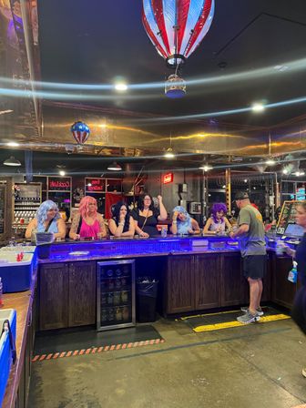 People in colorful wigs sitting at a blue LED-lit bar counter under red, white and blue hot-air balloon ceiling decorations as a bartender serves drinks in a lively pub interior.