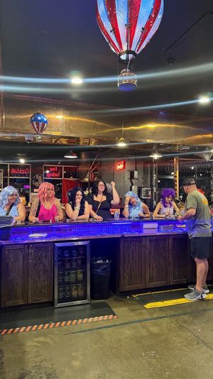 People in colorful wigs sitting at a blue LED-lit bar counter under red, white and blue hot-air balloon ceiling decorations as a bartender serves drinks in a lively pub interior.