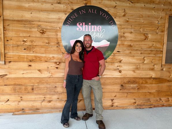 Smiling couple posing arm-in-arm in front of a round pink-and-black motivational sign on a rustic wood-paneled wall, wearing casual outfits and leopard-print shoes