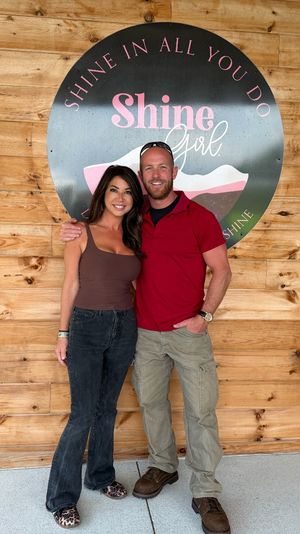 Smiling couple posing arm-in-arm in front of a round pink-and-black motivational sign on a rustic wood-paneled wall, wearing casual outfits and leopard-print shoes
