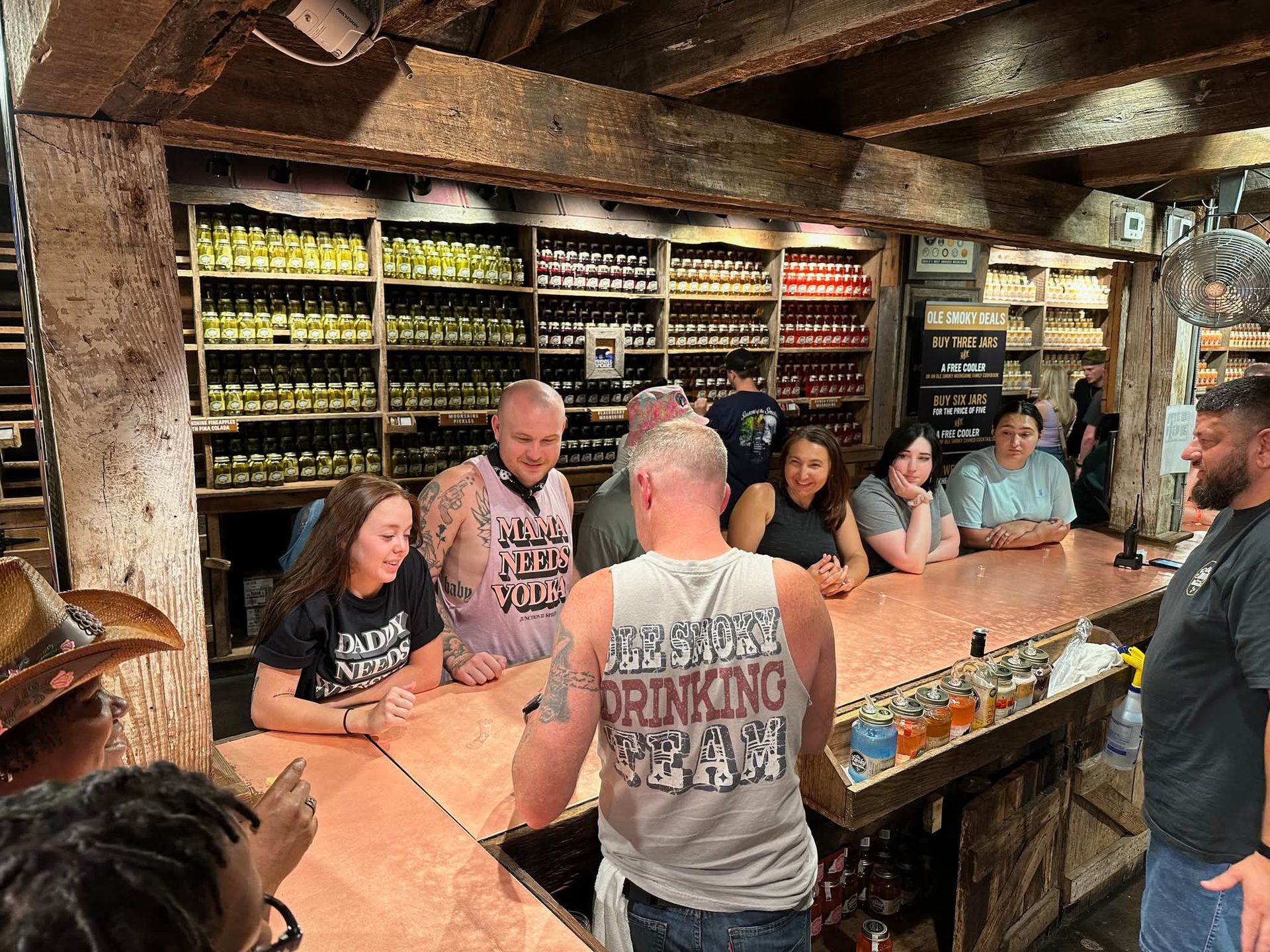 Bustling rustic tasting bar in a mountain‑town shop — people sampling colorful jarred spirits and preserves on wooden shelves beneath exposed beams
