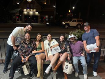 Seven friends posing on a bench outside a lit downtown main-street storefront at night, smiling and holding takeout boxes and shopping bags, several wearing cowboy boots.