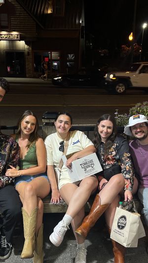 Seven friends posing on a bench outside a lit downtown main-street storefront at night, smiling and holding takeout boxes and shopping bags, several wearing cowboy boots.