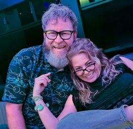 Smiling couple wearing glasses cuddling and laughing at an indoor lounge with blue-purple lighting; man with a beard in a patterned shirt and woman with wavy hair and bracelets on a casual night out.