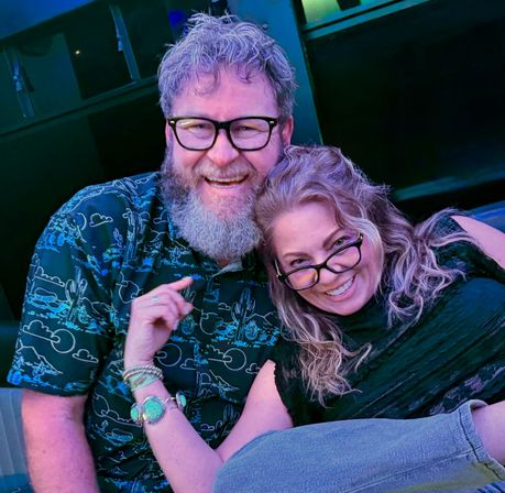 Smiling couple wearing glasses cuddling and laughing at an indoor lounge with blue-purple lighting; man with a beard in a patterned shirt and woman with wavy hair and bracelets on a casual night out.