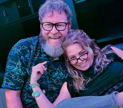 Smiling couple wearing glasses cuddling and laughing at an indoor lounge with blue-purple lighting; man with a beard in a patterned shirt and woman with wavy hair and bracelets on a casual night out.