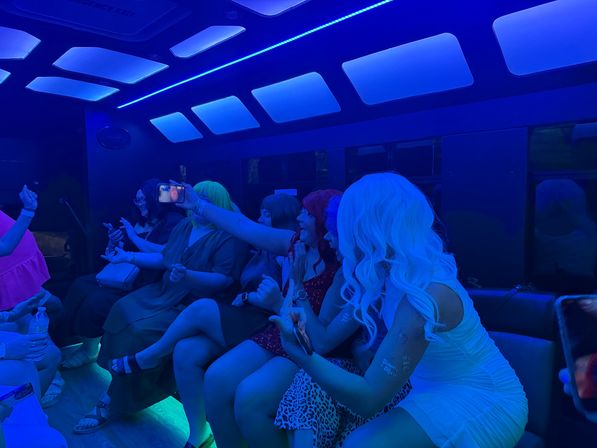 Friends partying on a blue-lit party bus, taking selfies and laughing in colorful wigs and dresses under neon ceiling panels