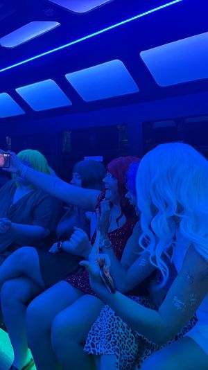 Friends partying on a blue-lit party bus, taking selfies and laughing in colorful wigs and dresses under neon ceiling panels