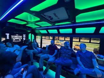 Glowing green-and-blue LED party shuttle interior with bench seating and a group of people chatting, windows showing parked cars and a grassy roadside.