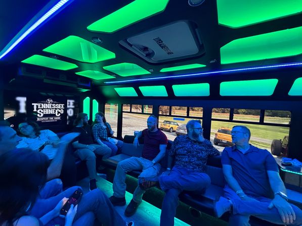 Glowing green-and-blue LED party shuttle interior with bench seating and a group of people chatting, windows showing parked cars and a grassy roadside.