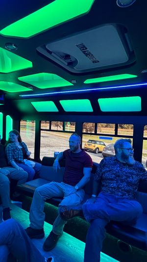 Glowing green-and-blue LED party shuttle interior with bench seating and a group of people chatting, windows showing parked cars and a grassy roadside.