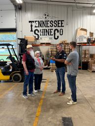Four people chatting in a Tennessee distillery warehouse beside stacked wooden barrels, industrial shelving and a parked forklift.