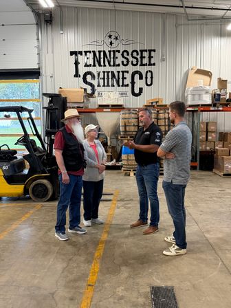 Four people chatting in a Tennessee distillery warehouse beside stacked wooden barrels, industrial shelving and a parked forklift.