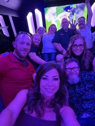 Smiling group selfie of nine adults inside a purple neon-lit party bus, friends laughing, waving and enjoying a nighttime celebration.