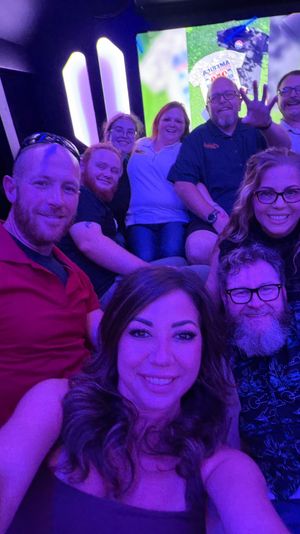 Smiling group selfie of nine adults inside a purple neon-lit party bus, friends laughing, waving and enjoying a nighttime celebration.