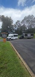 Small-town distillery parking lot with a wooden sign, a white passenger shuttle and a black delivery truck parked by tall trees under a bright, partly cloudy sky.