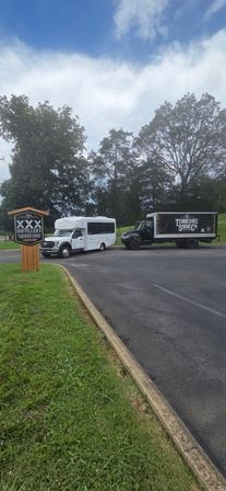 Small-town distillery parking lot with a wooden sign, a white passenger shuttle and a black delivery truck parked by tall trees under a bright, partly cloudy sky.