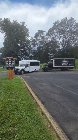 Small-town distillery parking lot with a wooden sign, a white passenger shuttle and a black delivery truck parked by tall trees under a bright, partly cloudy sky.