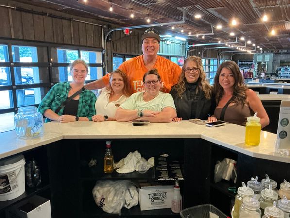 Six smiling adults posing at a rustic Tennessee tasting-room counter with jars of samples, a mason-jar drink, string lights and garage-style windows
