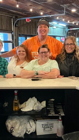Six smiling adults posing at a rustic Tennessee tasting-room counter with jars of samples, a mason-jar drink, string lights and garage-style windows