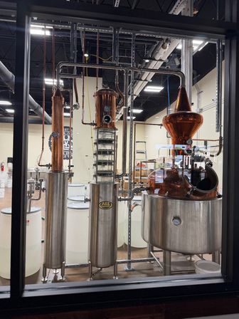 Gleaming copper and stainless-steel distillation stills behind a glass window in a craft distillery, featuring a pot still with onion-shaped copper head, tall reflux column, interconnected piping and white fermentation tanks.
