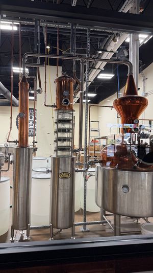 Gleaming copper and stainless-steel distillation stills behind a glass window in a craft distillery, featuring a pot still with onion-shaped copper head, tall reflux column, interconnected piping and white fermentation tanks.