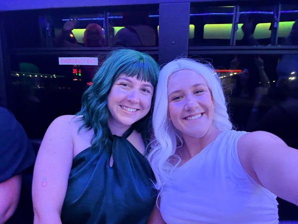 Two smiling people with teal and white hair taking a selfie under blue-purple party lights inside a party shuttle, city nightlife vibe