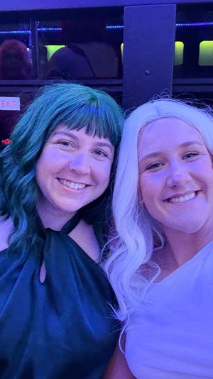 Two smiling people with teal and white hair taking a selfie under blue-purple party lights inside a party shuttle, city nightlife vibe