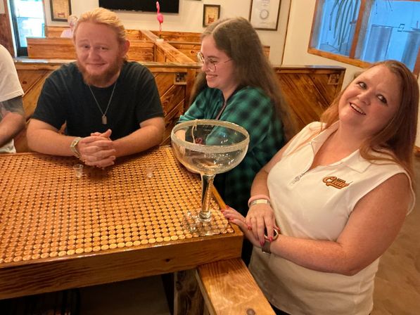 Three smiling patrons at a cozy wooden bar seated at a penny-covered tabletop with a giant margarita-style glass bowl