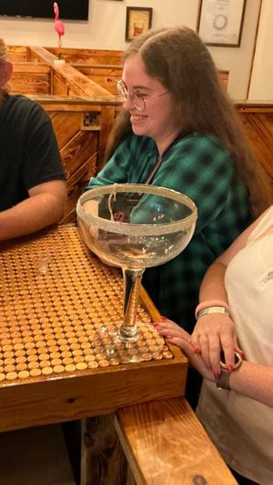 Three smiling patrons at a cozy wooden bar seated at a penny-covered tabletop with a giant margarita-style glass bowl