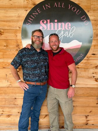 Two smiling men with arms around each other pose in front of a large round decorative sign mounted on a wood-paneled wall; one wears a patterned button-up, jeans and glasses with a beard, the other wears a red polo, cargo pants and sunglasses on his head.