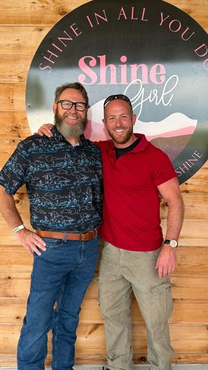 Two smiling men with arms around each other pose in front of a large round decorative sign mounted on a wood-paneled wall; one wears a patterned button-up, jeans and glasses with a beard, the other wears a red polo, cargo pants and sunglasses on his head.