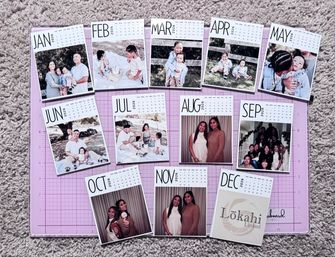 Flat lay of a custom 2026 photo calendar — twelve month cards (JAN–DEC) with family and portrait photos arranged on a pink craft cutting mat over beige carpet.