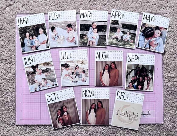 Flat lay of a custom 2026 photo calendar — twelve month cards (JAN–DEC) with family and portrait photos arranged on a pink craft cutting mat over beige carpet.