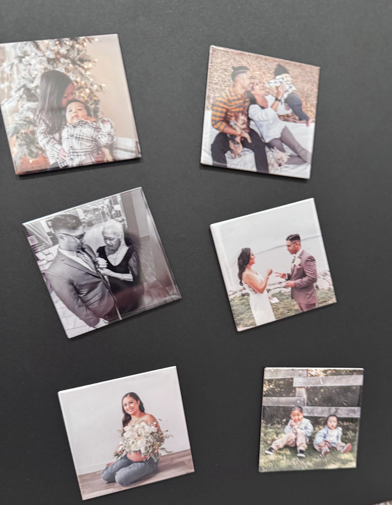 Seven square custom photo magnets on a black background showing family scenes: holiday mom with infant, parents with baby and pets, grayscale groom with older woman, wedding couple exchanging rings, pregnant woman with bouquet, and two toddlers outdoors.
