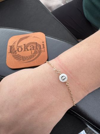 Close-up of an ankle wearing a delicate gold chain anklet with a white round initial 'D' charm, resting on a black leather surface beside a brown leather coaster stamped with a circular logo.