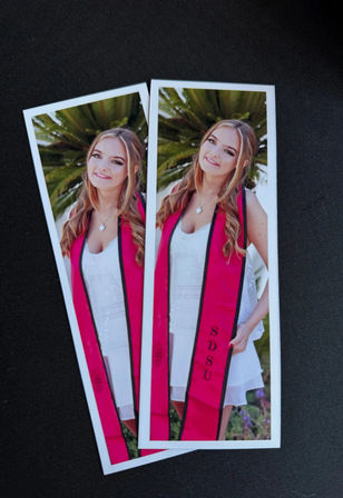Two long photo prints of a smiling graduate in a white dress wearing a bright pink graduation sash with black trim, palm fronds background on a black textured surface.