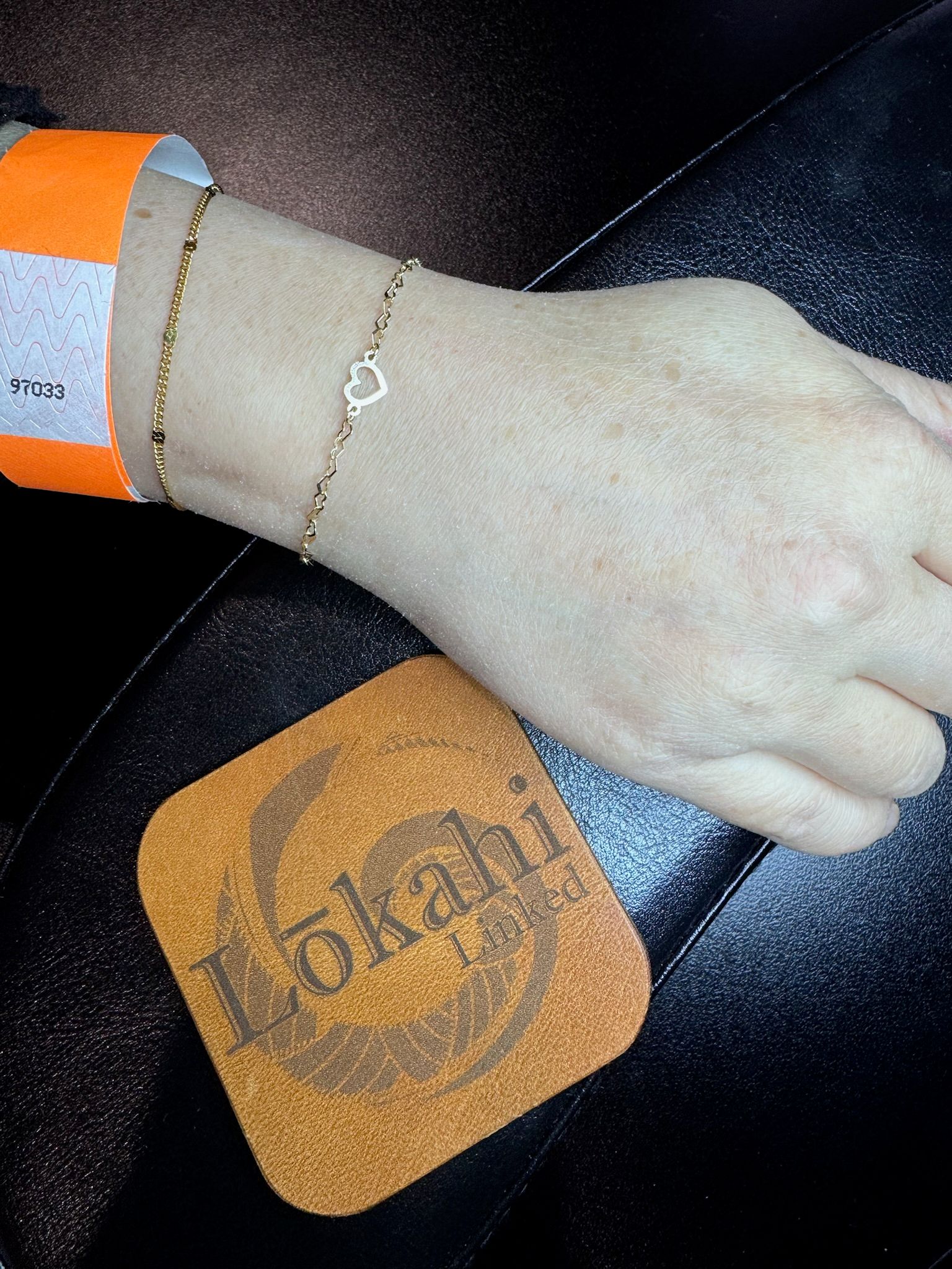 Close-up of a wrist wearing an orange paper wristband and two delicate gold bracelets — one with a tiny open-heart charm — resting on a black leather surface beside a tan leather coaster.