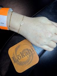 Close-up of a wrist wearing an orange paper wristband and two delicate gold bracelets — one with a tiny open-heart charm — resting on a black leather surface beside a tan leather coaster.
