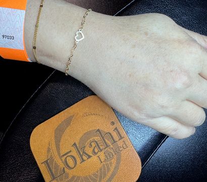 Close-up of a wrist wearing an orange paper wristband and two delicate gold bracelets — one with a tiny open-heart charm — resting on a black leather surface beside a tan leather coaster.