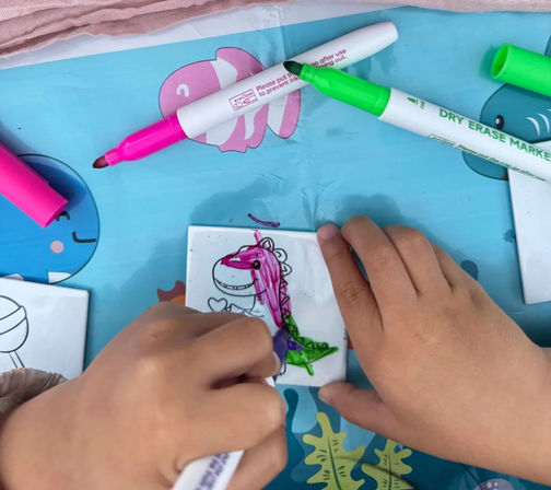 Kids' hands coloring a cartoon dinosaur with pink and green dry-erase markers on a blue play mat, soft pink fabric draped above.
